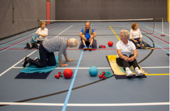 Deelnemers zitten op een matje in de gymzaal met blauwe grond.