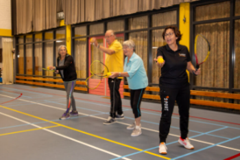 Een docent en drie deelnemers die met een tennisracket een bal proberen weg te slaan.